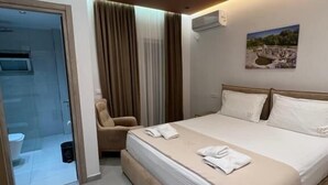 Habitació d'hotel moderna amb un llit gran, una cadira, un bany amb porta de vidre i un quadre a la paret.