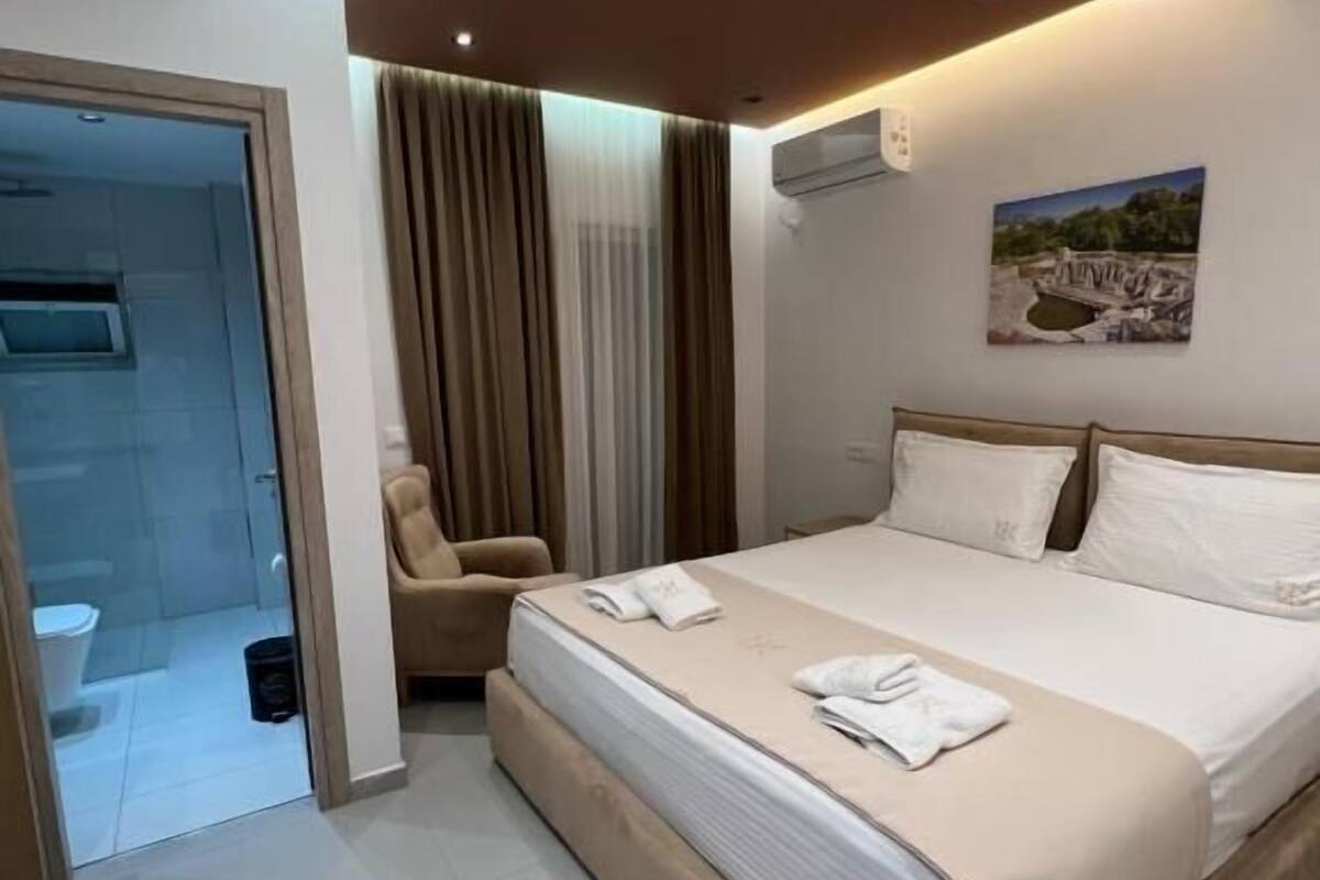 A modern hotel room with a large bed, a chair, a bathroom with a glass door, and a painting on the wall.