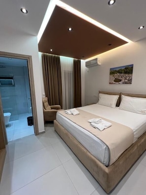 A modern hotel room with a large bed, a chair, a bathroom with a glass door, and a painting on the wall. - KRONOS Hotel Sarande (Sarande)