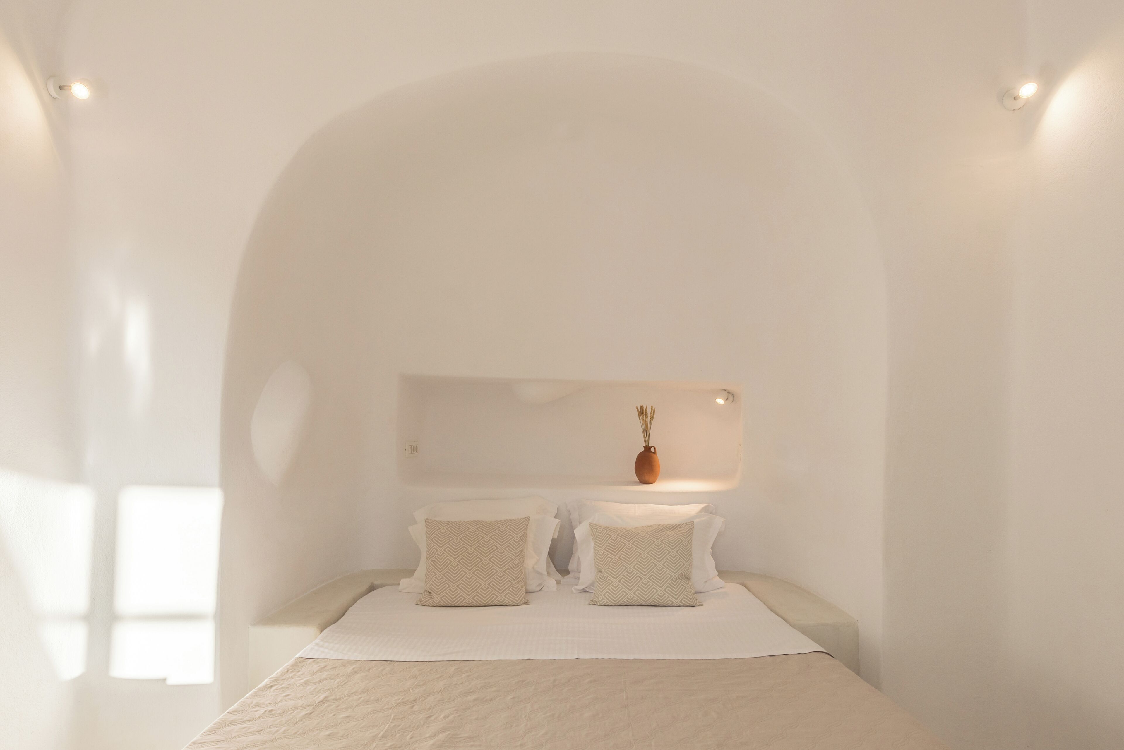 A minimalist bedroom with a low arched ceiling, a bed with two pillows, a small vase on the wall, and a single light fixture.