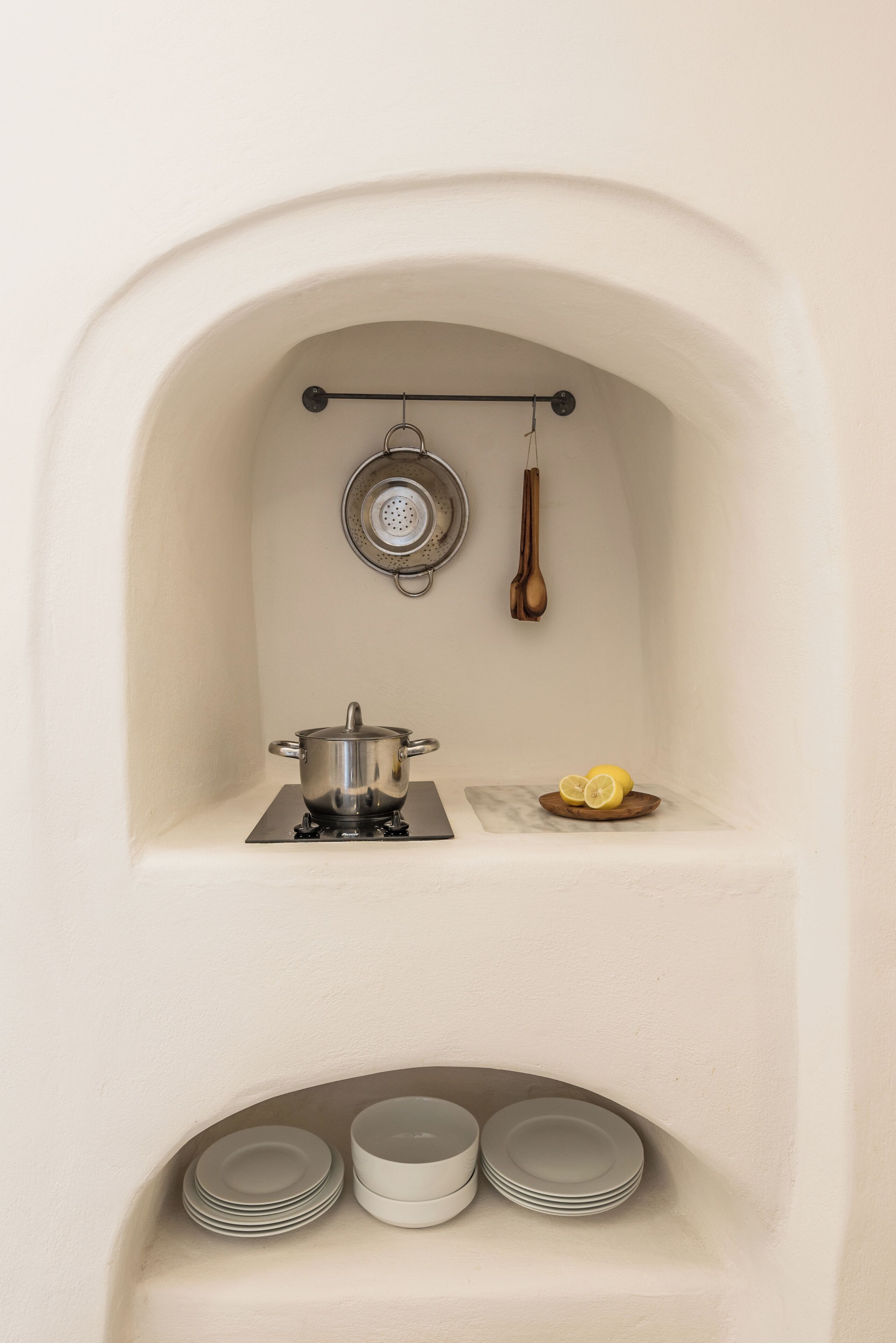 Una kitchenette pequeña con estufa, ollas, colador, utensilios de madera y platos apilados.