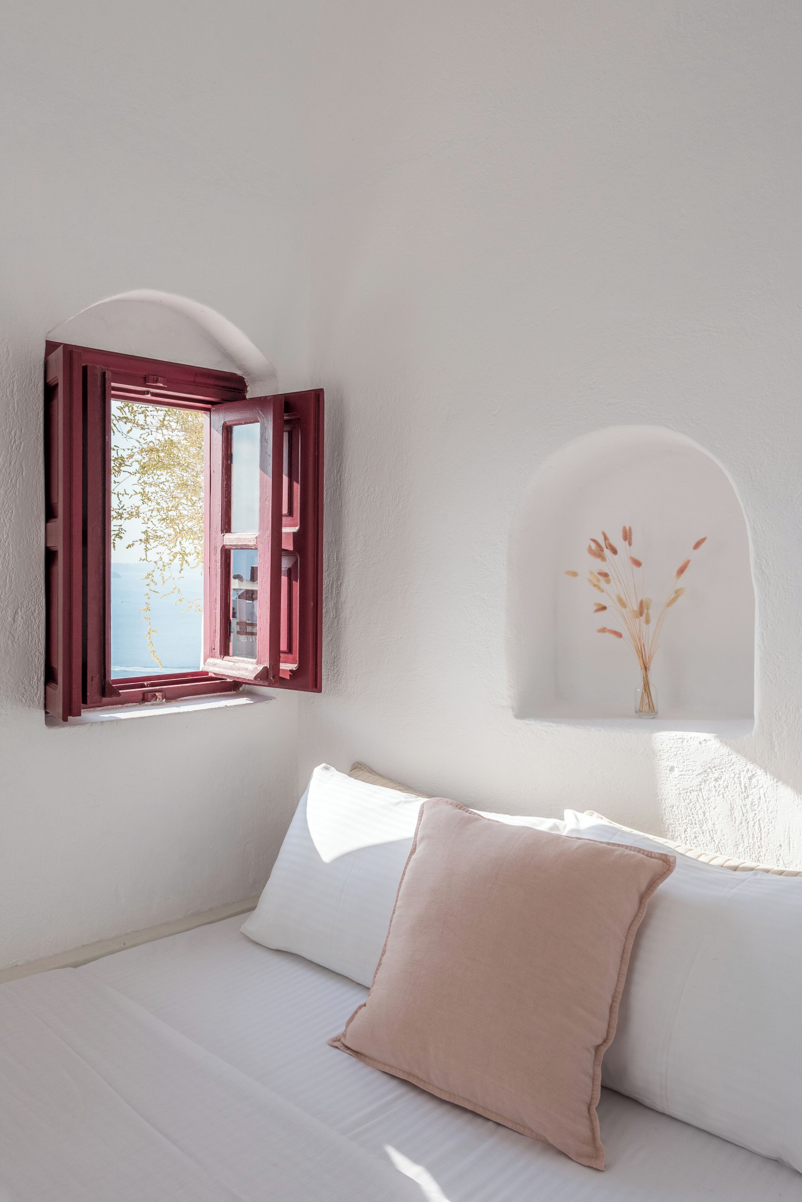 A room with a bed, a window with red shutters, a vase with dried flowers, and a white wall.