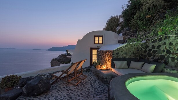 A seaside villa with a stone wall, a wooden chair, a sofa, and a hot tub. - Cycladica Santorini (Oia)