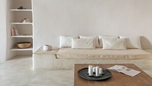 A minimalist living room with a beige sofa, white cushions, a wooden coffee table with candles and a magazine, and a white shelving unit with books and a basket. - Cycladica Santorini (Oia)