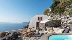 A stone-floored outdoor area with a pool, a red door, and a white building with a view of the sea. - Cycladica Santorini (Oia)