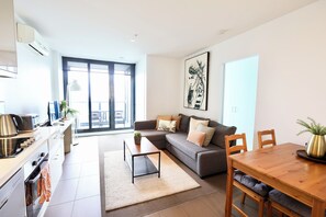 A modern living room with a sofa, a dining table, and a kitchen area with a stove and sink. - Beautiful Harbour View Apartment + Free Car Park (Melbourne)