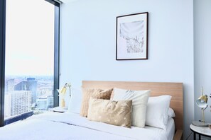 Une chambre d’hôtel avec une grande fenêtre, un lit avec de nombreux oreillers, un tableau encadré au mur et une table de chevet avec une lampe.