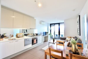 A modern kitchen with white cabinets, a dining area with a wooden table, and a living room with a sofa and a large window. - Beautiful Harbour View Apartment + Free Car Park (Melbourne)