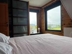 A bedroom with a bed, a nightstand, a lamp, and a window with a view of the outdoors. - Villa with access to lapin plage, view of the marsh, not overlooked. Restful. (La Hague)