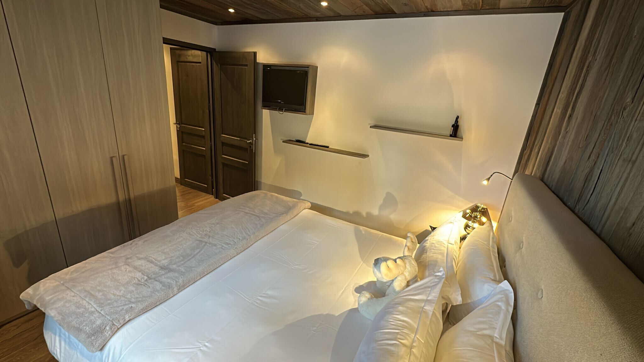 A bedroom with a bed, a TV mounted on the wall, a shelf, and a wardrobe.