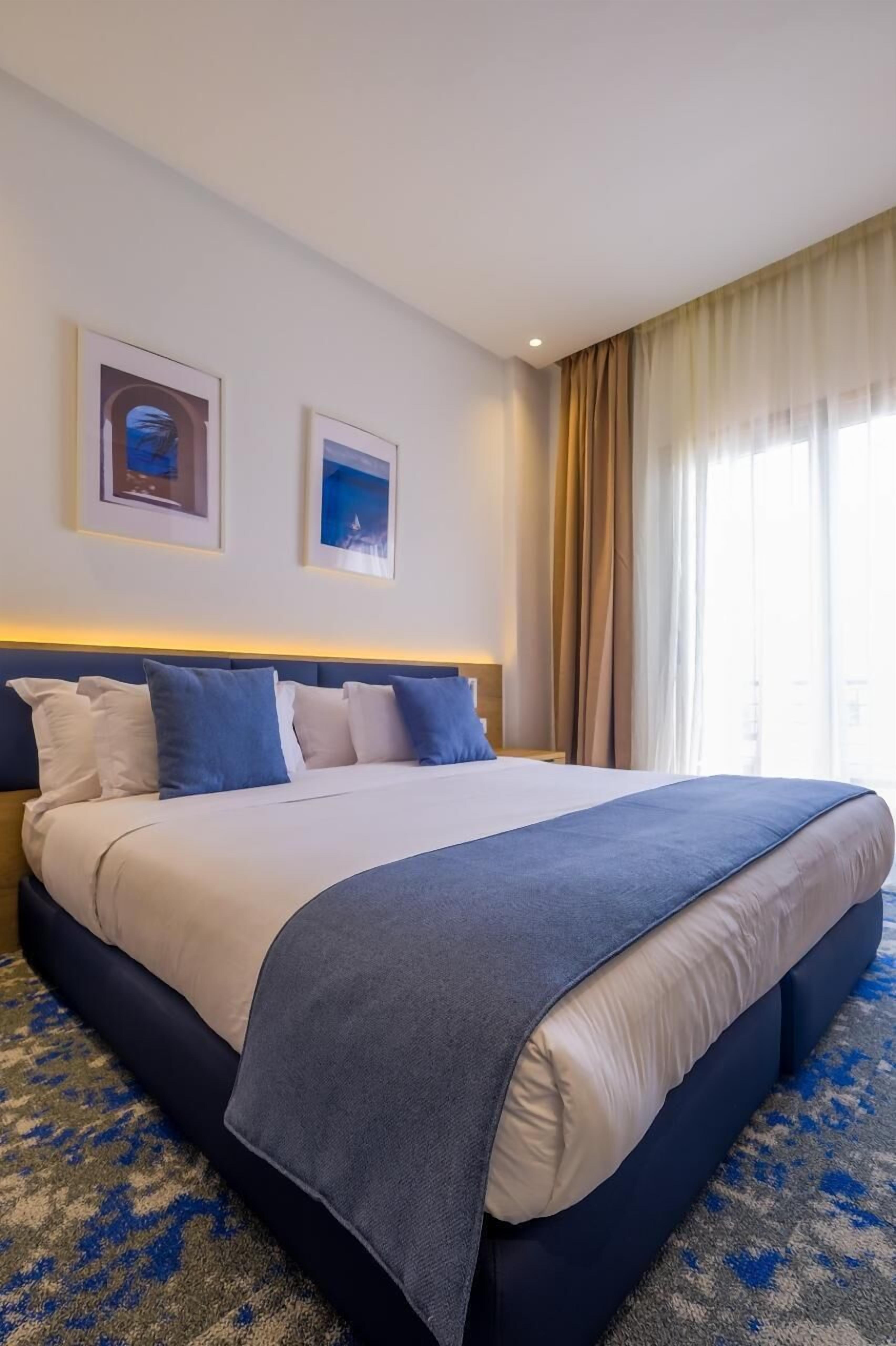 A hotel room with a large bed, a headboard, two framed pictures on the wall, and a window with curtains.