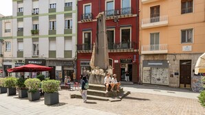 Una plaza pública con un monumento central, edificios circundantes con tiendas y mesas al aire libre.