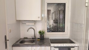 A compact kitchen with white cabinets, a stainless steel sink, a stove, and a small window with a decorative view. - SEASIDE STUDIO (Calcatoggio)