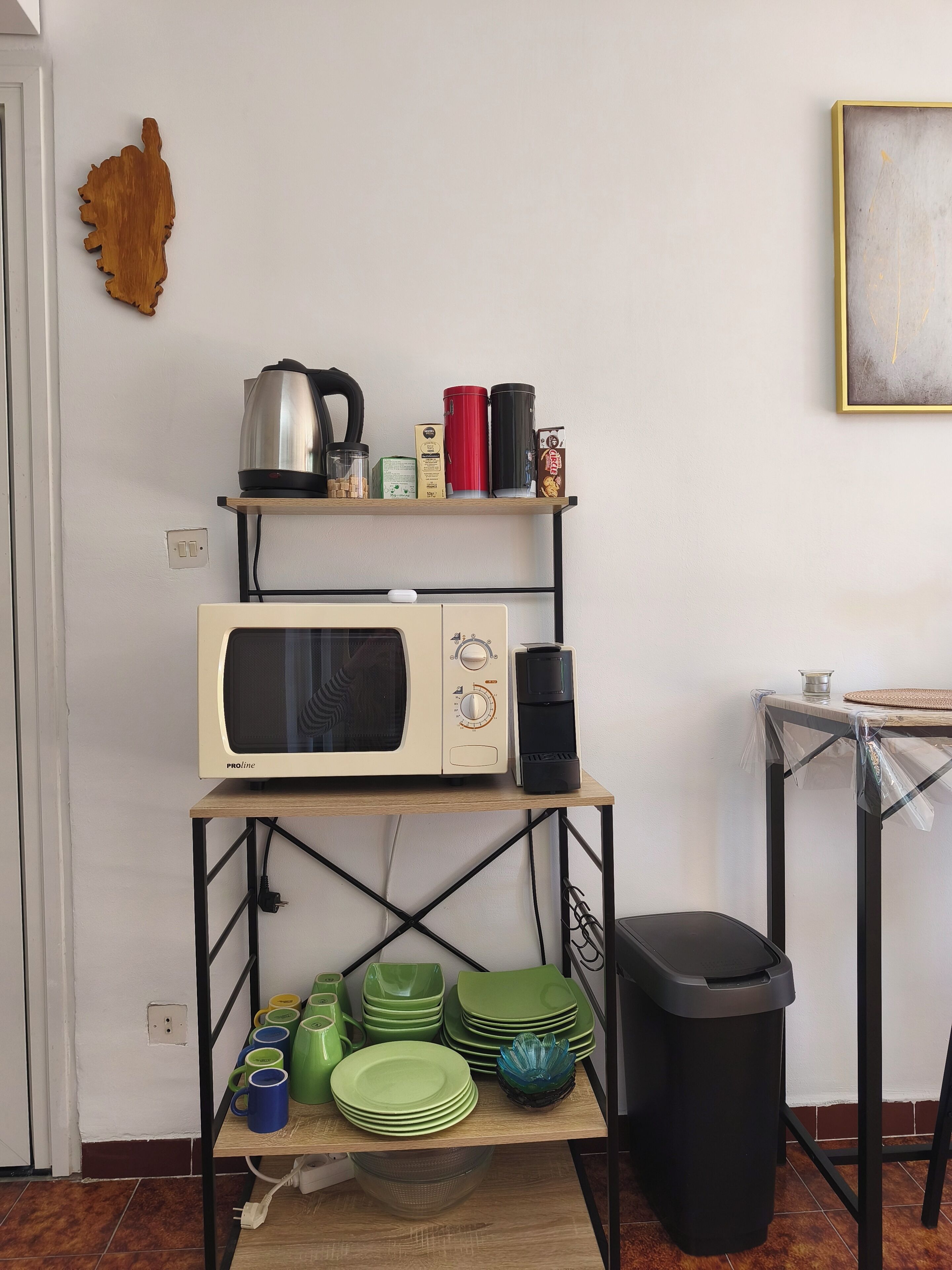 A kitchen with a microwave, shelves with kitchenware, a trash bin, and a framed picture on the wall.