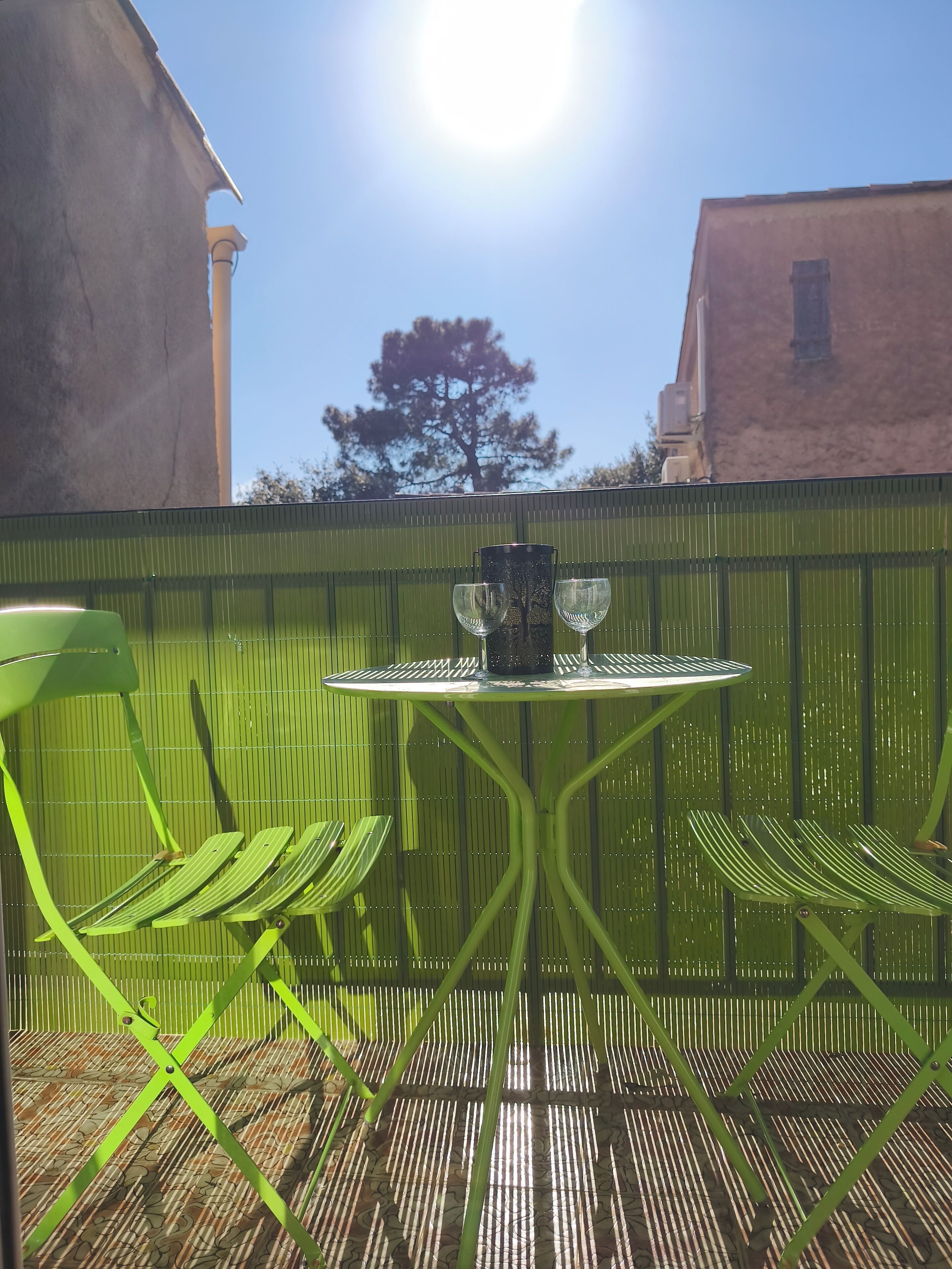 Une table en métal verte, avec deux chaises et un verre dessus, placée devant une clôture verte, avec un arbre en arrière-plan.