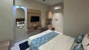 A hotel room with a bed, a TV, a mirror, a ceiling fan, a small kitchenette, and a bathroom. - Hotel Prime Elite (Ahmedabad)