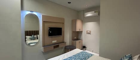 Habitación de hotel con cama, televisión, espejo, ventilador de techo, kitchenette pequeña y baño.