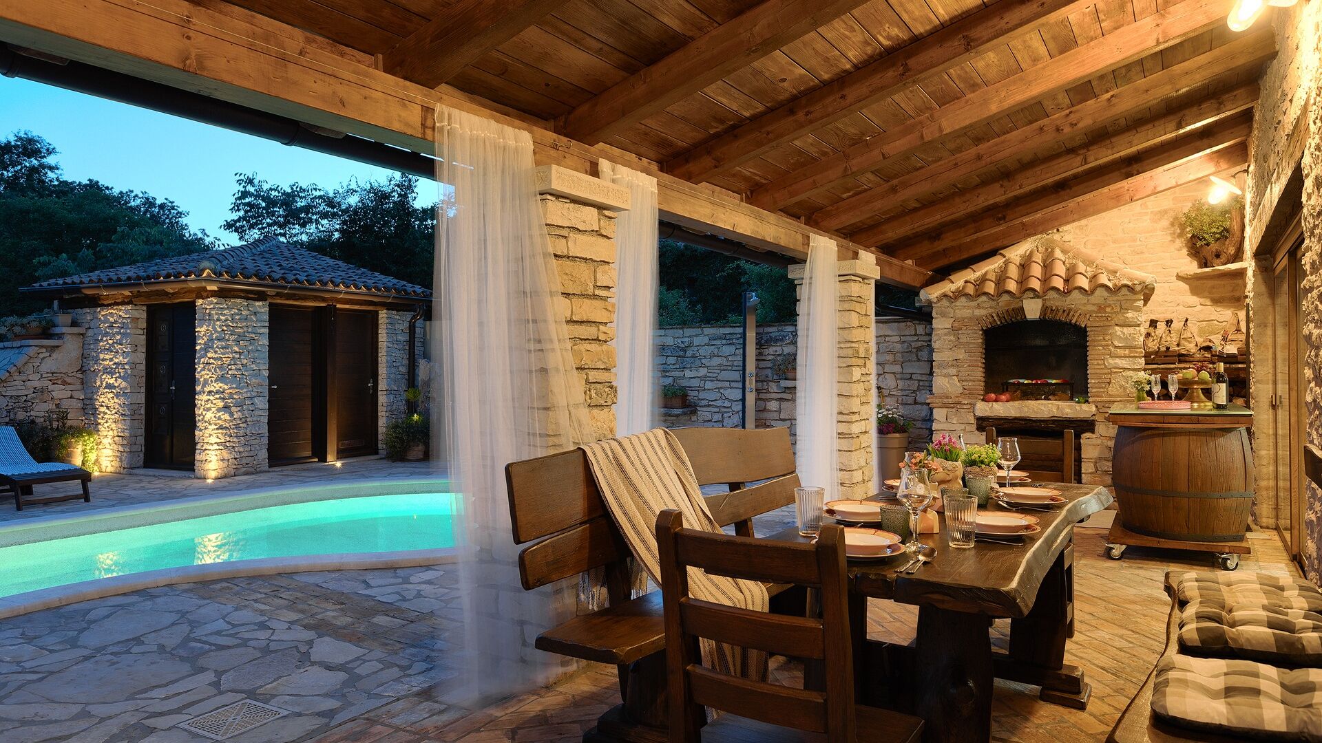 An outdoor patio with a pool, stone walls, wooden furniture, and a stone fireplace.
