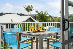 Une table en bois, des chaises et une carafe en verre sont disposées sur un balcon doté d’une balustrade blanche.