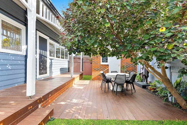A backyard with a wooden deck, a round table with chairs, and a tree providing shade. - Rathay House - Victoria Park. Close to CBD, Optus stadium, Swan River, Crown. (Victoria Park)