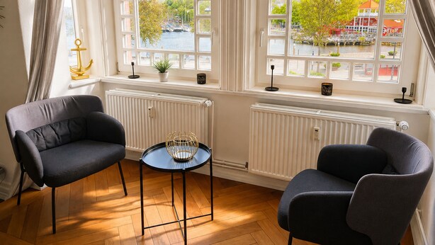 A room with two armchairs, a small table, and a view of a river and buildings through large windows. - fewo1846 - Strandkoje / luxurious old apartment for max 5 persons with sea view (Flensburg)