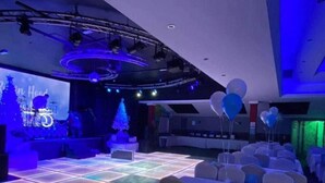 Un salón de banquetes con mesas dispuestas para un evento, un escenario con batería y un árbol de Navidad.