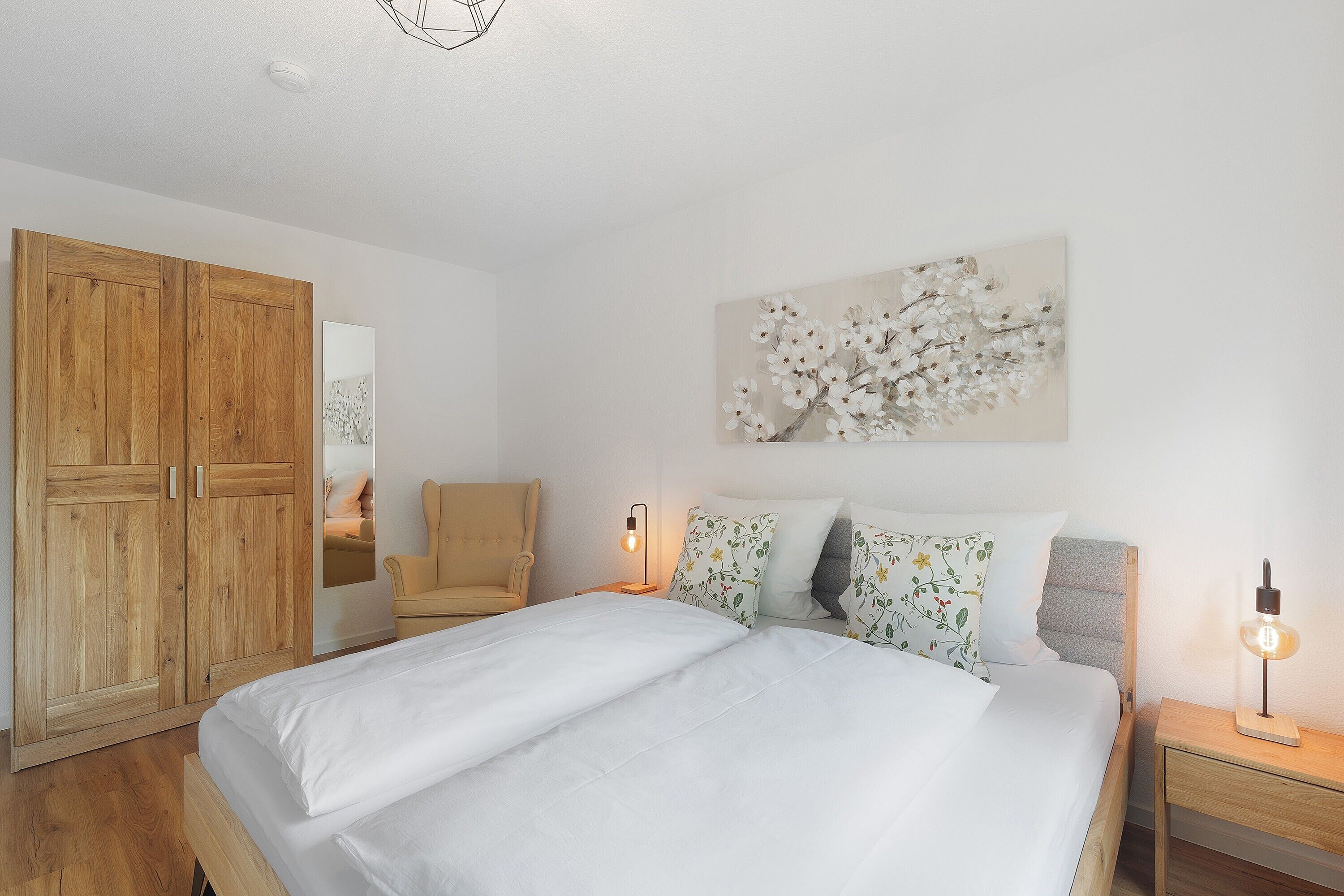 A bedroom with a bed, a wooden wardrobe, a chair, a bedside table with a lamp, and a wall-mounted artwork.