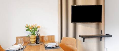 Un comedor con una mesa de madera, sillas, un televisor montado en la pared y un pequeño estante.