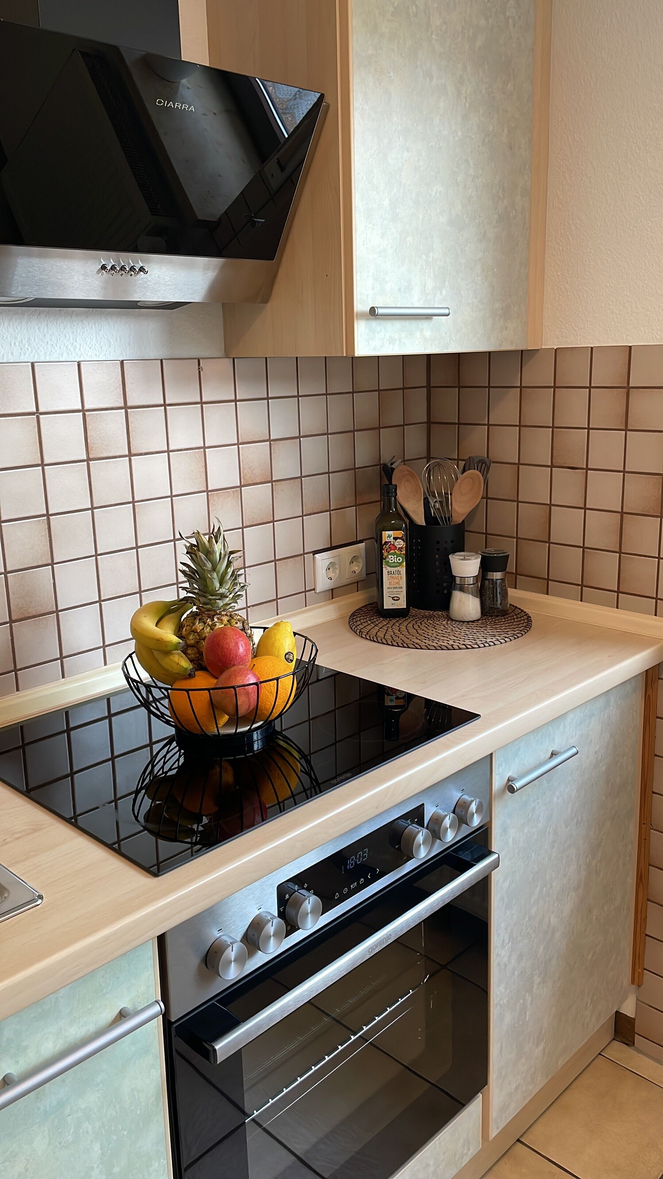 Una cocina con una placa de inducción negra, un horno empotrado y un microondas. Hay un frutero en la encimera y una botella de aceite de oliva.