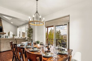 Ein Essbereich mit einem Holztisch, gedeckt für vier Personen, umgeben von Stühlen, einer Kristallleuchtre und Blick ins Freie durch große Fenster.