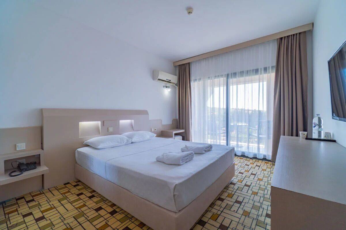 Habitación de hotel con cama, mesitas de noche, unidad de aire acondicionado, un ventanal con cortinas y televisor de pantalla plana.
