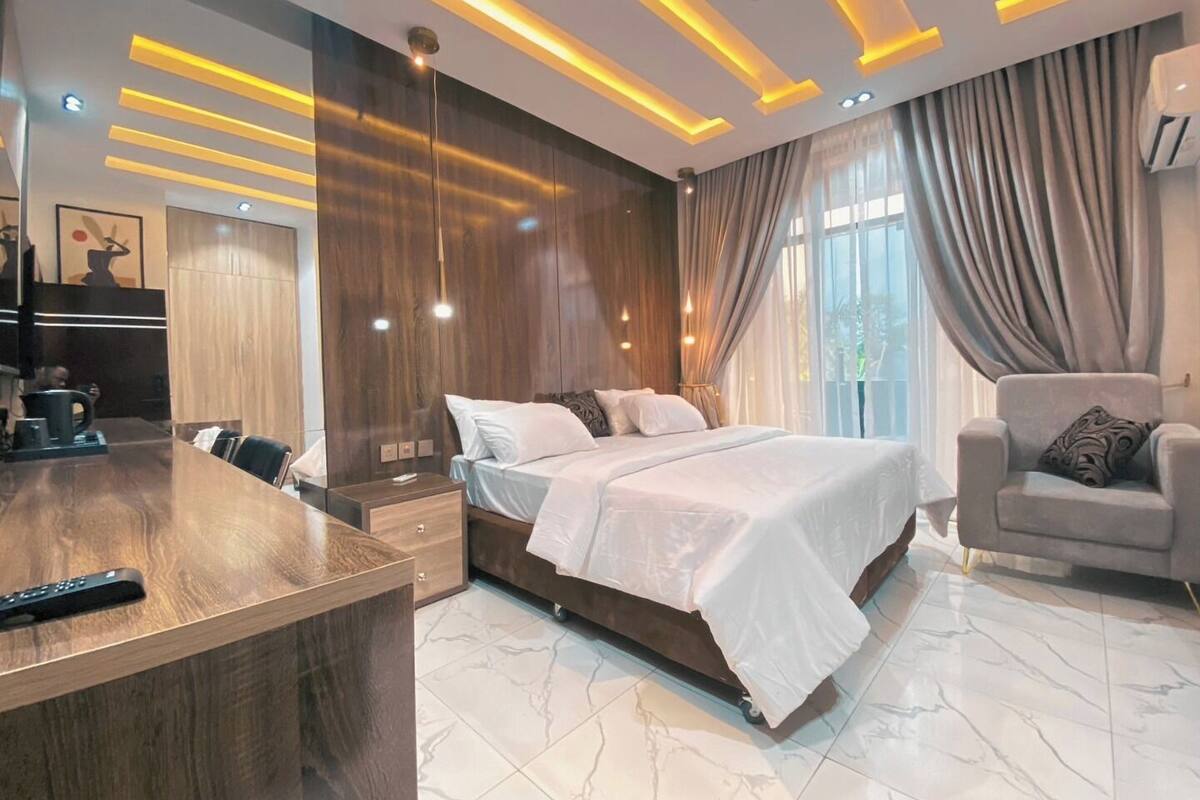 Ein modernes Hotelzimmer mit einem Holztisch, einem großen Bett, einem grauen Sessel und einem großen Fenster mit Vorhängen.