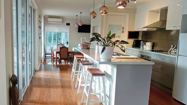 A modern kitchen with a central island, bar stools, and a dining area with chairs. - 3BR Bushland Retreat, Relax/ Professional Stay–near the bay & CBD/ Private Deck (North Batemans Bay)