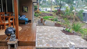 A wooden deck with a stone pathway leading to a seating area with a blue sofa and wooden chairs. - 3BR Bushland Retreat, Relax/ Professional Stay–near the bay & CBD/ Private Deck (North Batemans Bay)