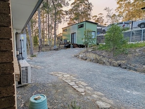 Un camino de grava conduce a una casa moderna con una pequeña terraza, un depósito de agua y un generador eléctrico.