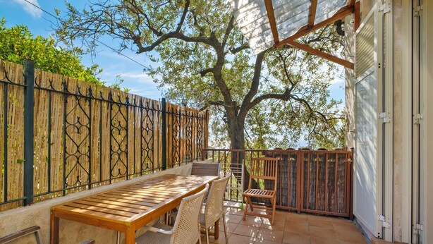 Een terras buiten met een houten tafel en stoelen, een houten omheining en een boom die schaduw geeft.