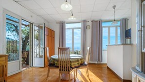 Een ruime kamer met een houten vloer, een ronde tafel met stoelen, een groot raam met gordijnen en een televisie aan de muur.