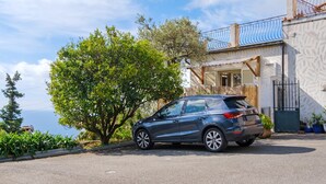 Een blauwe SUV geparkeerd voor een huis met een balkon, een boom en uitzicht op zee.