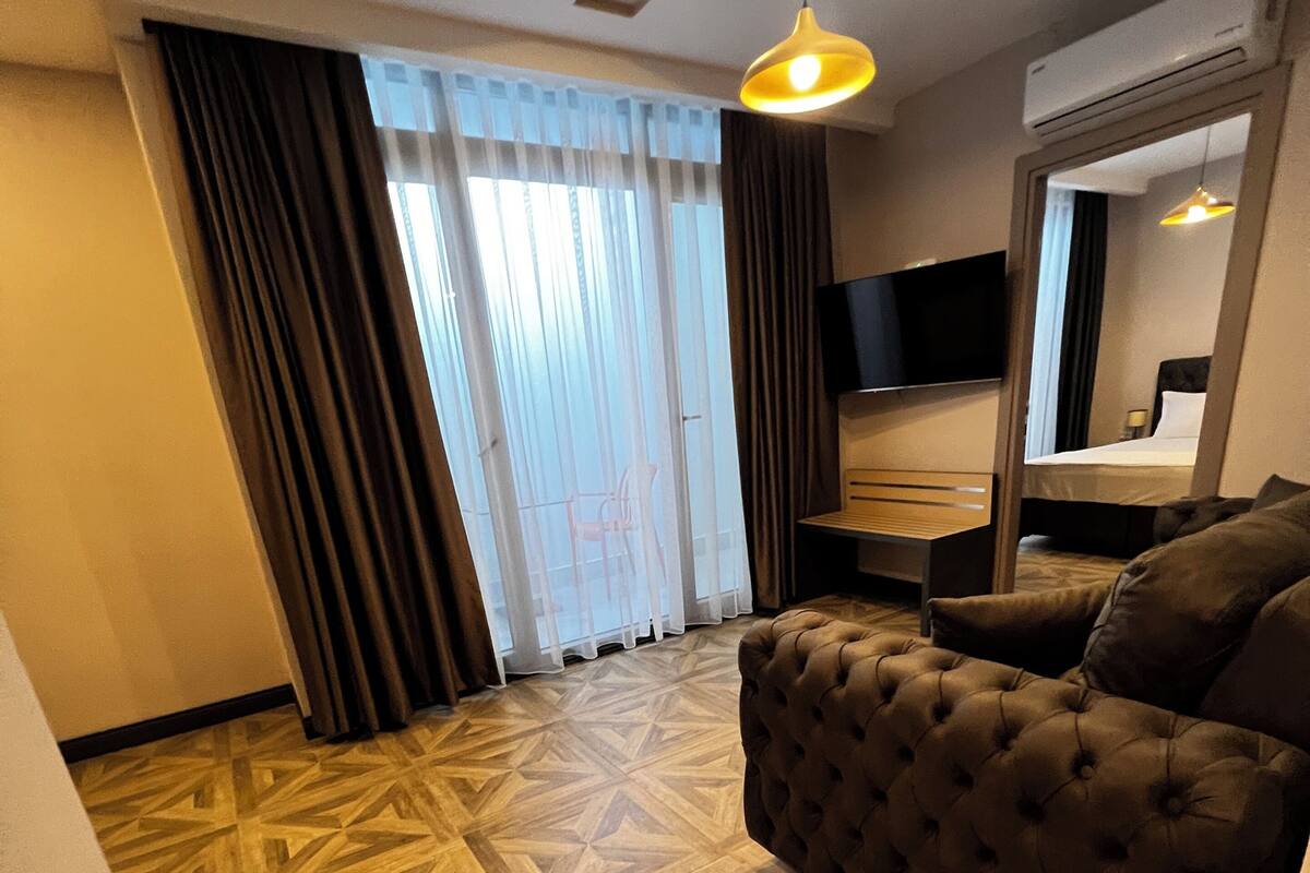 A hotel room with a patterned wooden floor, a tufted sofa, a television, a bed, and a large window with curtains.