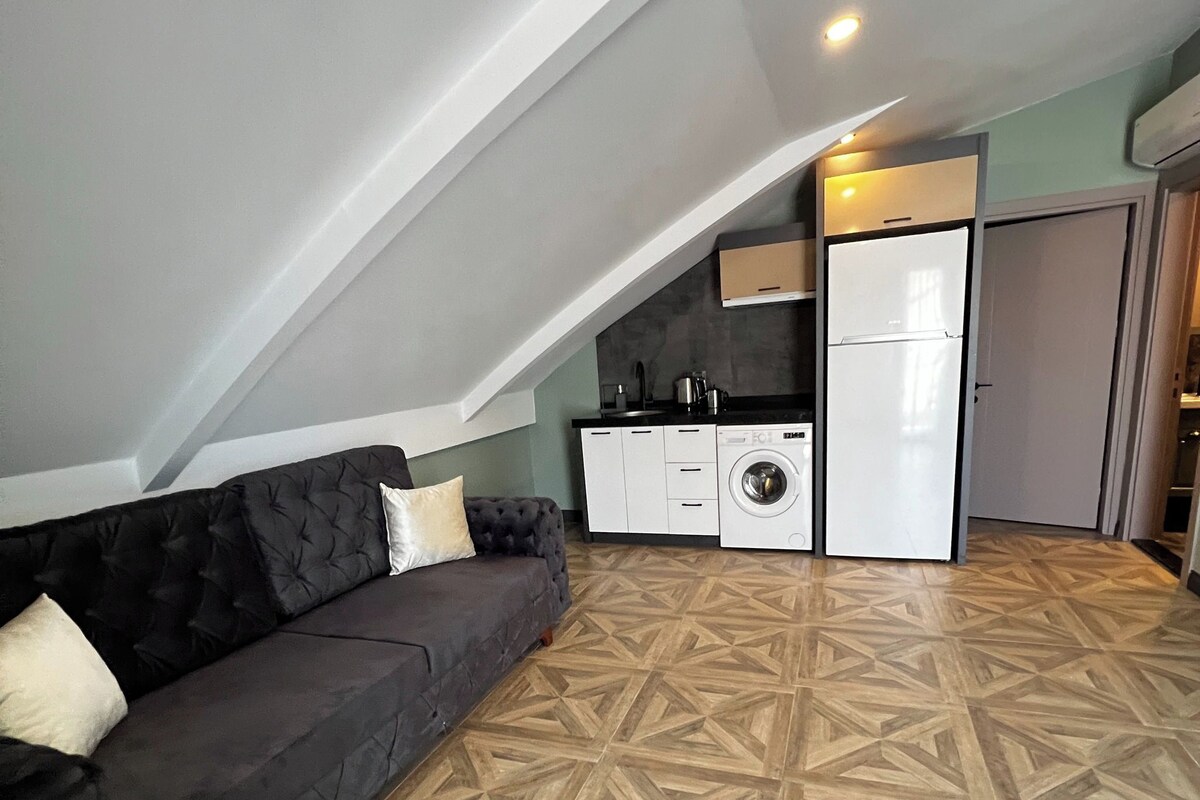 A loft-style living space with a dark sofa, a washing machine, and a refrigerator.