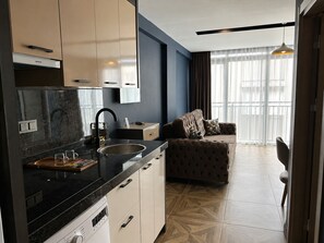 Una cocina moderna con encimera negra, un lavaplatos integrado y un lavabo. Cuenta con una zona de estar con sofá y sillón, además de un ventanal con cortinas.