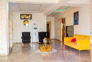 Un hall d’entrée avec un sol en marbre, deux fauteuils noirs, un canapé jaune, une table basse à cadre doré et un distributeur d’eau fixé au mur.