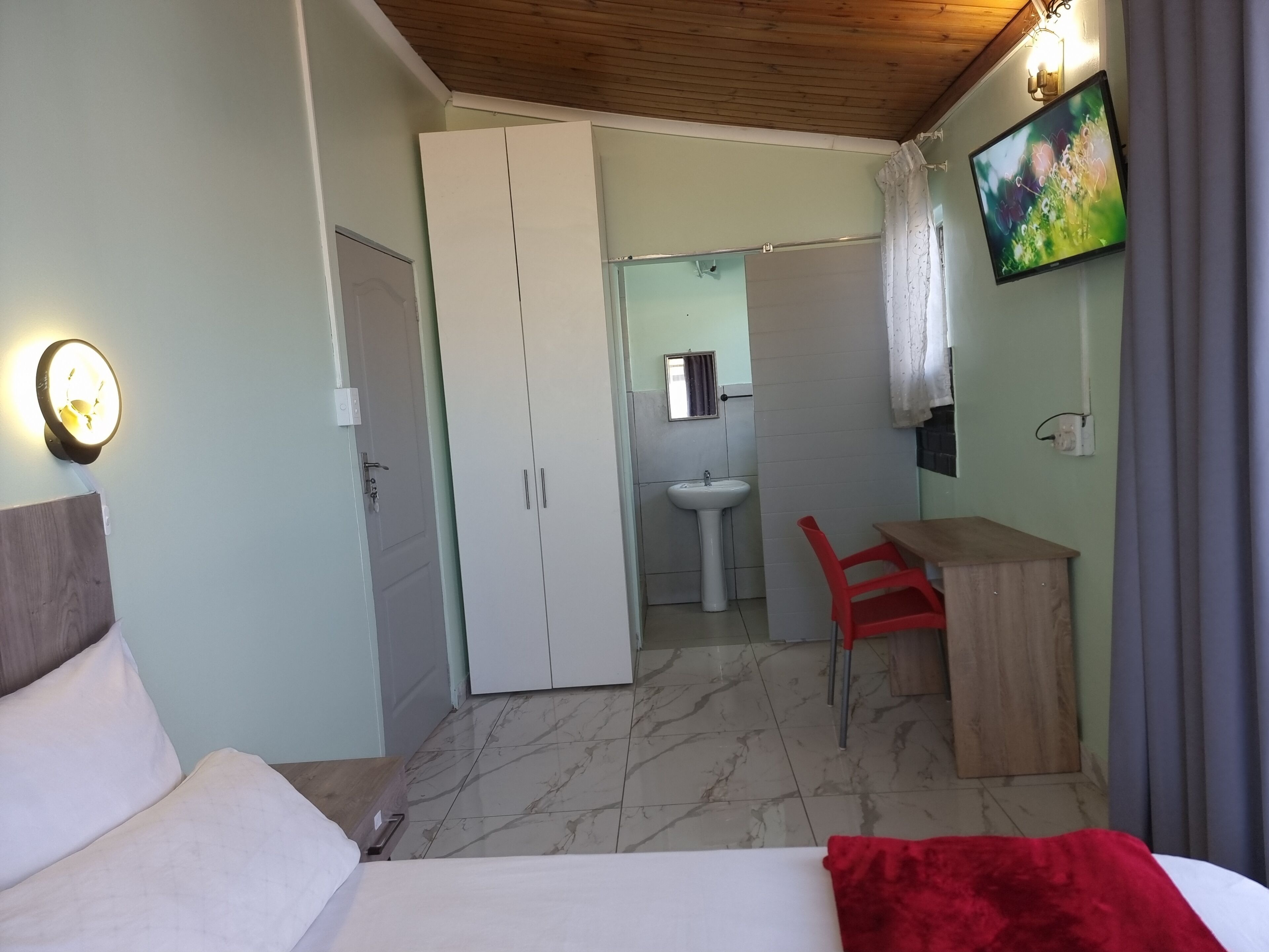 A bedroom with a bed, a desk with a chair, a wardrobe, a mirror, a sink, and a television.