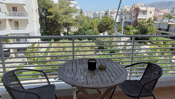 Ein Balkon mit rundem Tisch und zwei Stühlen, Blick auf Gebäude und Grünflächen, und ein klarer Himmel.