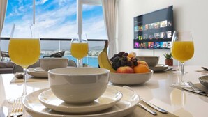 Une table à manger avec un saladier de fruits, deux verres de jus d’orange et un téléviseur diffusant un film.