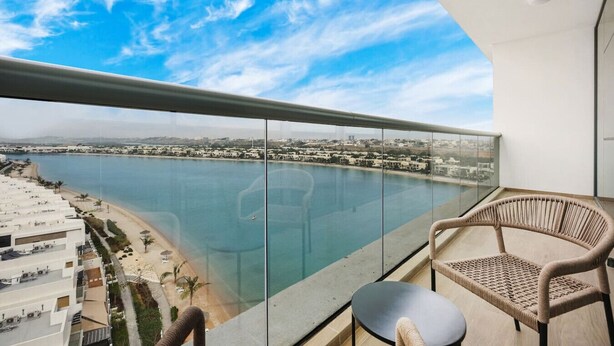 Un balcon doté d’une balustrade en verre, offrant une vue sur une étendue d’eau, une plage et le paysage urbain.