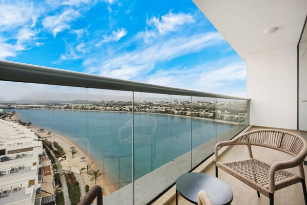 A balcony with a glass railing overlooking a body of water, a beach, and a cityscape. - FIRST CLASS |1BR| Sea View Paradise | Scenic Views (Ras Al Khaimah)