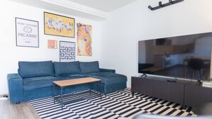 Ein modernes Wohnzimmer mit einem blauen Sofa, einem Holztisch, einem schwarz-weißen Streifen Teppich, einem Flachbildfernseher und Kunstwerken an der Wand.
