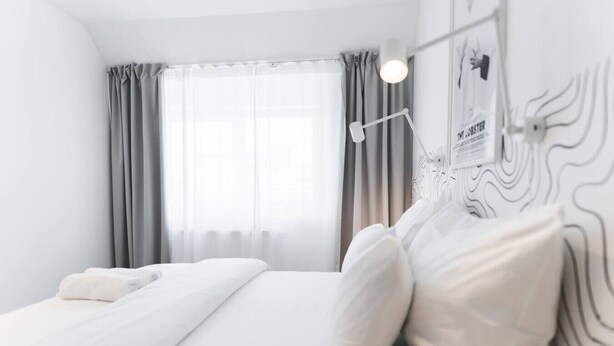 Ein modernes Hotelzimmer mit einem Bett, einem Nachttisch, einer Lampe und Blick ins Freie durch das Fenster.