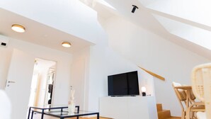 A modern loft apartment with a high ceiling, a flat-screen TV mounted on a white cabinet, a dining area with a glass table, and a wicker chair. - Two leveled Penthouse Schönbrunn (Vienna)